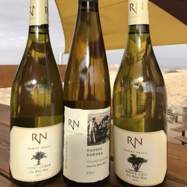 שלושה בקבוקי יין לבן של יקב רמת נגב ניצבים על בר העץ וברקע הנוף המדברי המוכר של חוות קורנמל