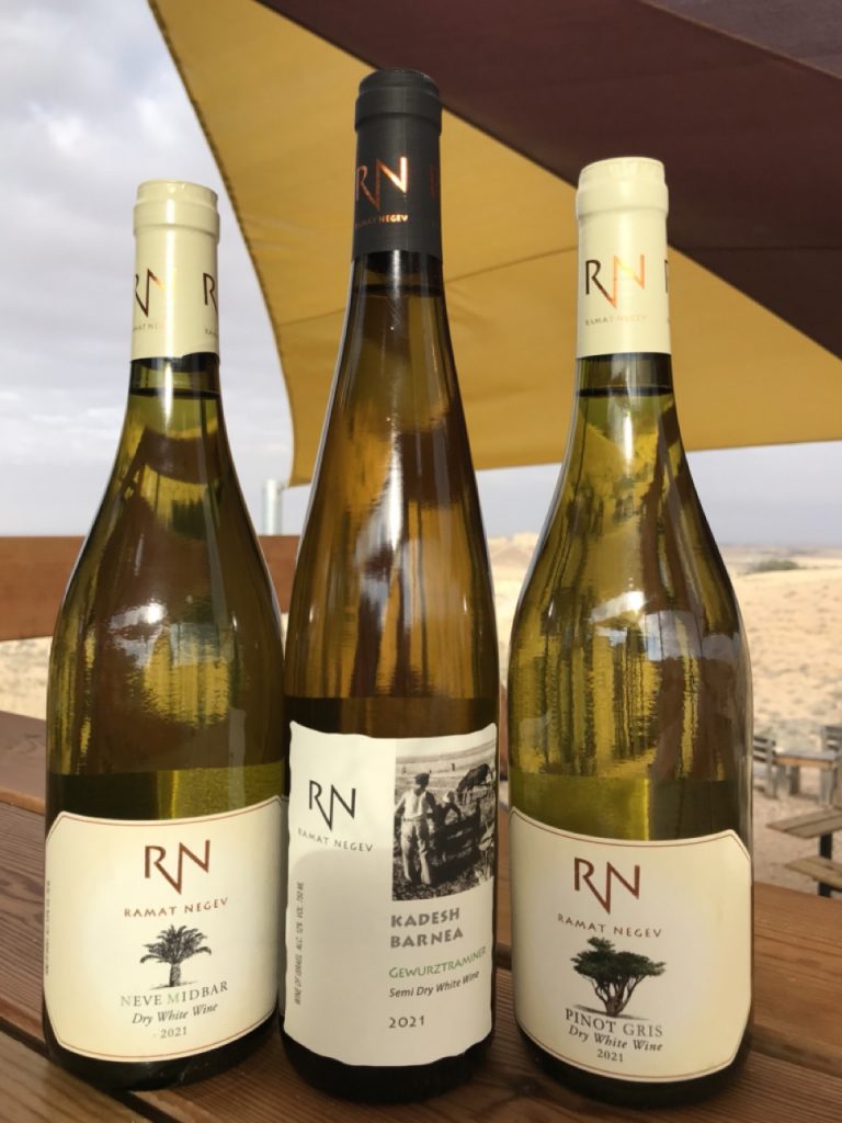 שלושה בקבוקי יין לבן של יקב רמת נגב ניצבים על בר העץ וברקע הנוף המדברי המוכר של חוות קורנמל