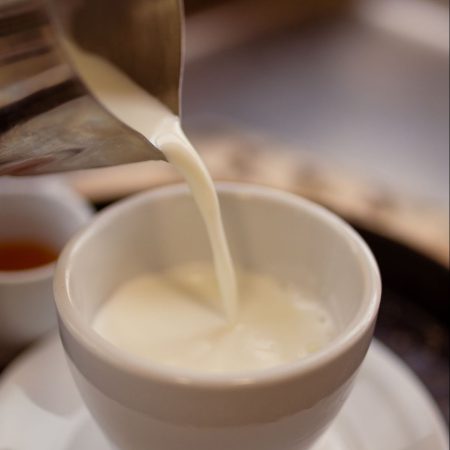 כד הקצפה מתכתי המוזג חלב חם לכוס חרסינה לבנה המונחת על צלוחית הגשה. מוגש עם כוסית דבש בצד.